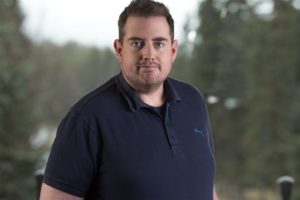 Man in a navy polo shirt standing indoors, with a blurred greenery background. he appears focused and is looking directly at the camera.