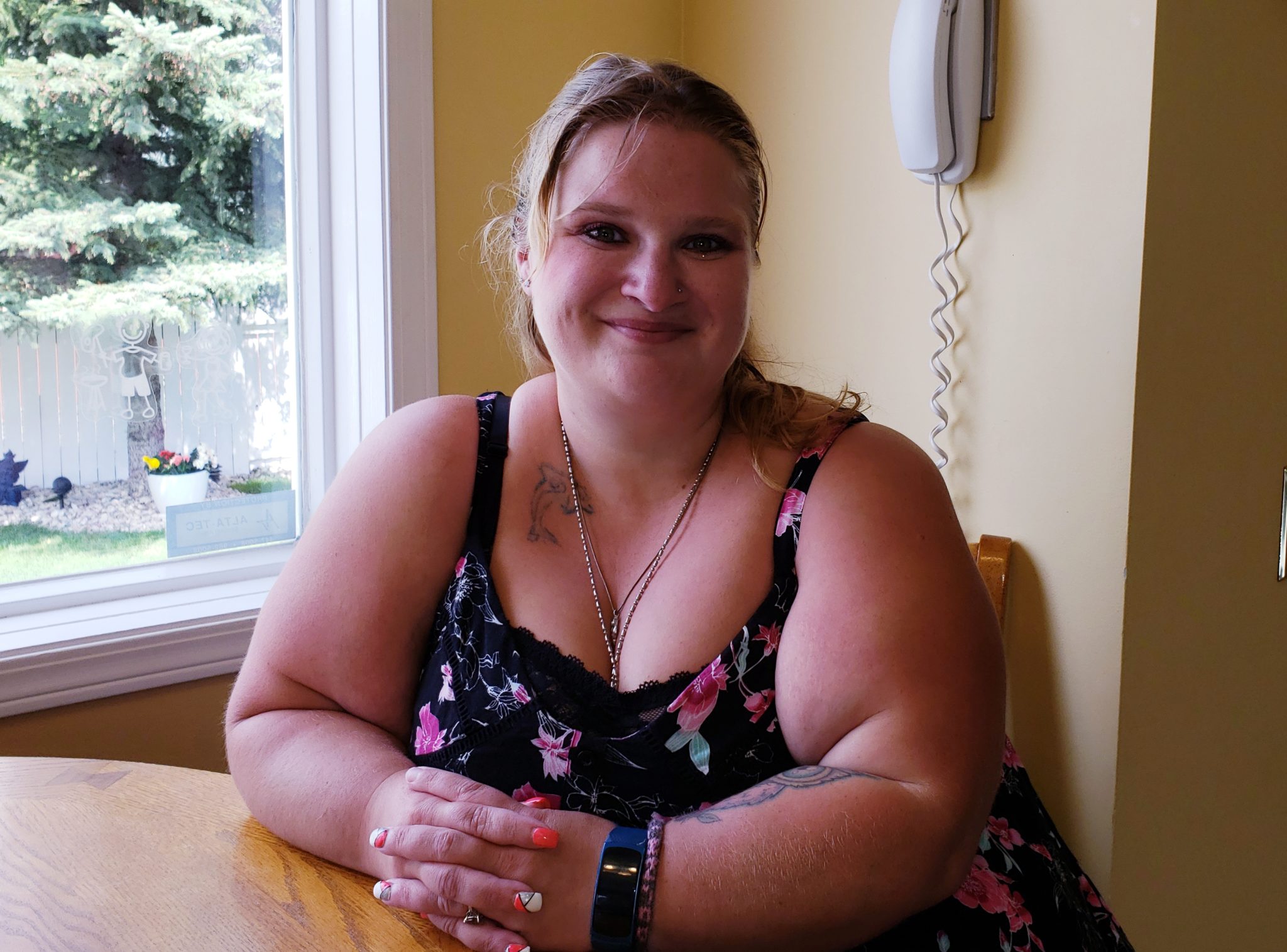 Woman sitting at a table indoors, smiling at the camera, with a garden visible through the window behind her. she wears a floral dress and a necklace.