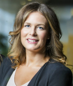 Professional woman with brown hair smiling, wearing a black blazer and white top, in a well-lit office setting.