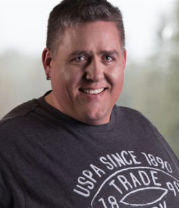 A portrait of a smiling man wearing a dark t-shirt with text, against a blurred background.