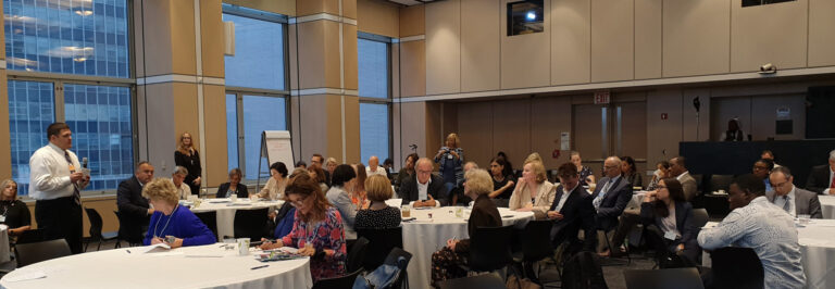 A man standing and speaking at a business conference with an attentive audience seated around tables in a room with large windows.