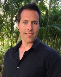 A man with dark hair wearing a black shirt smiles slightly, standing outdoors with palm trees in the background.