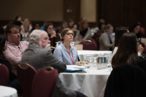 Audience members focused intently during a conference, seated at tables, with conference materials visible.