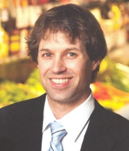 A smiling man with shaggy brown hair, wearing a dark suit and light blue tie, with a colorful blurred background.