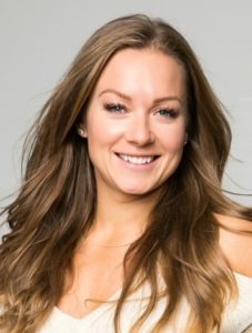 Portrait of a smiling woman with long brown hair and earrings, wearing a white top against a gray background.