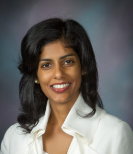 Professional headshot of a woman with dark hair and a white blazer, smiling against a blurred blue background.