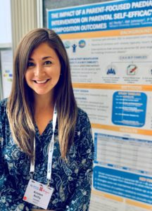 A young woman presenting a research poster at a conference, wearing a lanyard with a name tag and smiling at the camera.