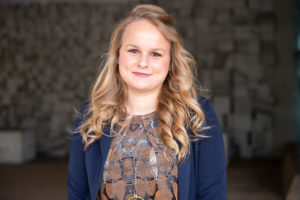 A woman with curly blonde hair smiling at the camera, wearing a blue blazer and a patterned blouse, standing in front of a textured wall.