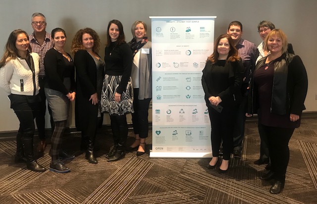 Group of eleven professionals, mixed gender, standing in a business meeting room alongside a presentation poster.