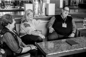 Three adults laughing and talking on a couch; two women and a man, all wearing conference name tags.