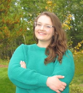 Woman in a teal sweater and glasses smiling with arms crossed, standing in front of autumn trees.