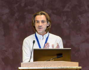 A man with shoulder-length hair speaking at a podium with a laptop, wearing a blue lanyard, against a textured brown background.