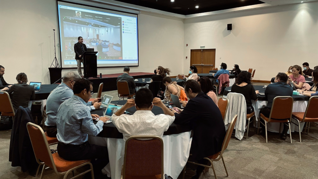 A conference setting with attendees seated at tables, focusing on laptops, while a speaker presents on the stage in front of a large screen.