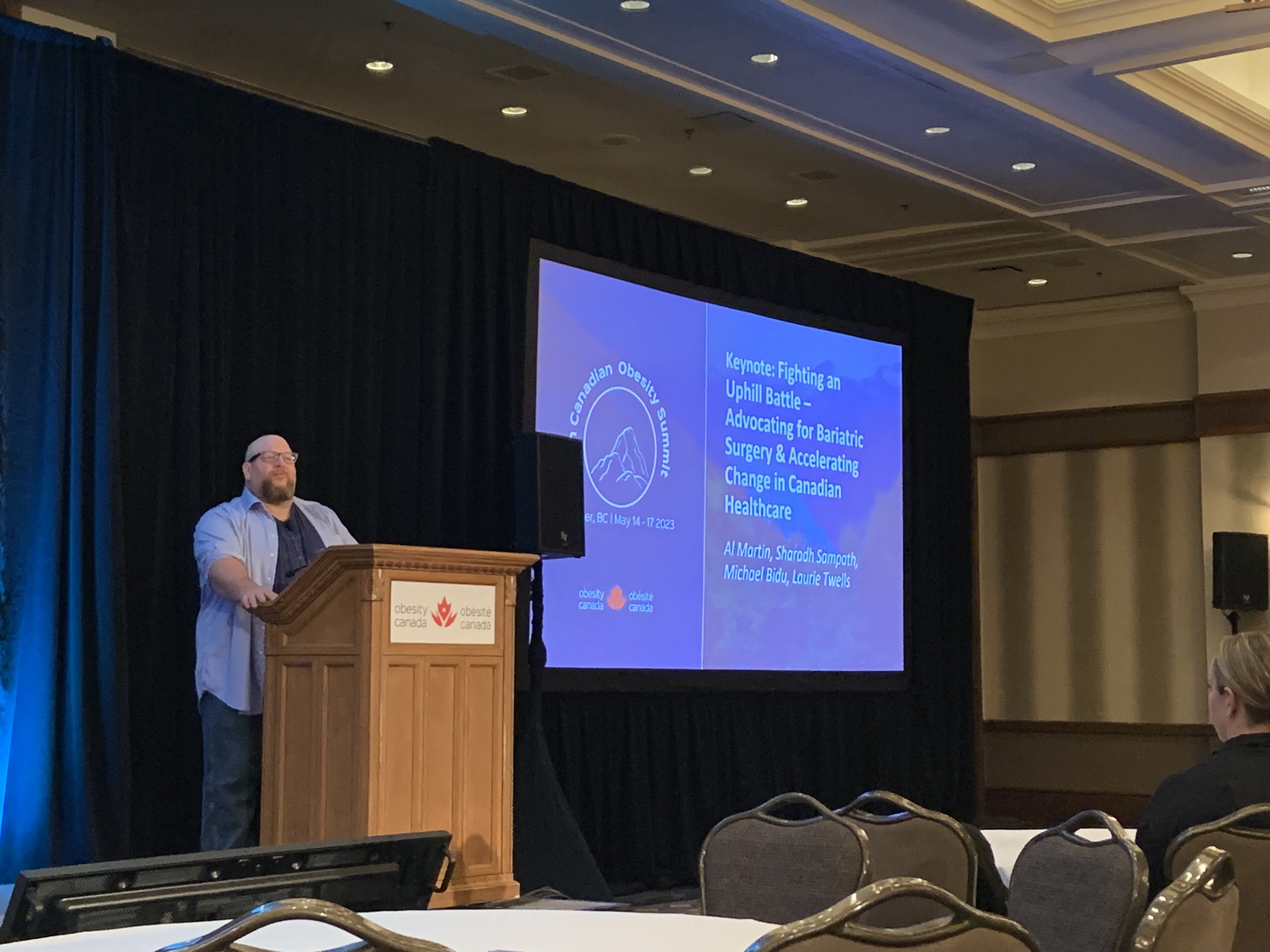 Man presenting at a conference with a slide about bariatric surgery advocacy in canadian healthcare projected behind him.