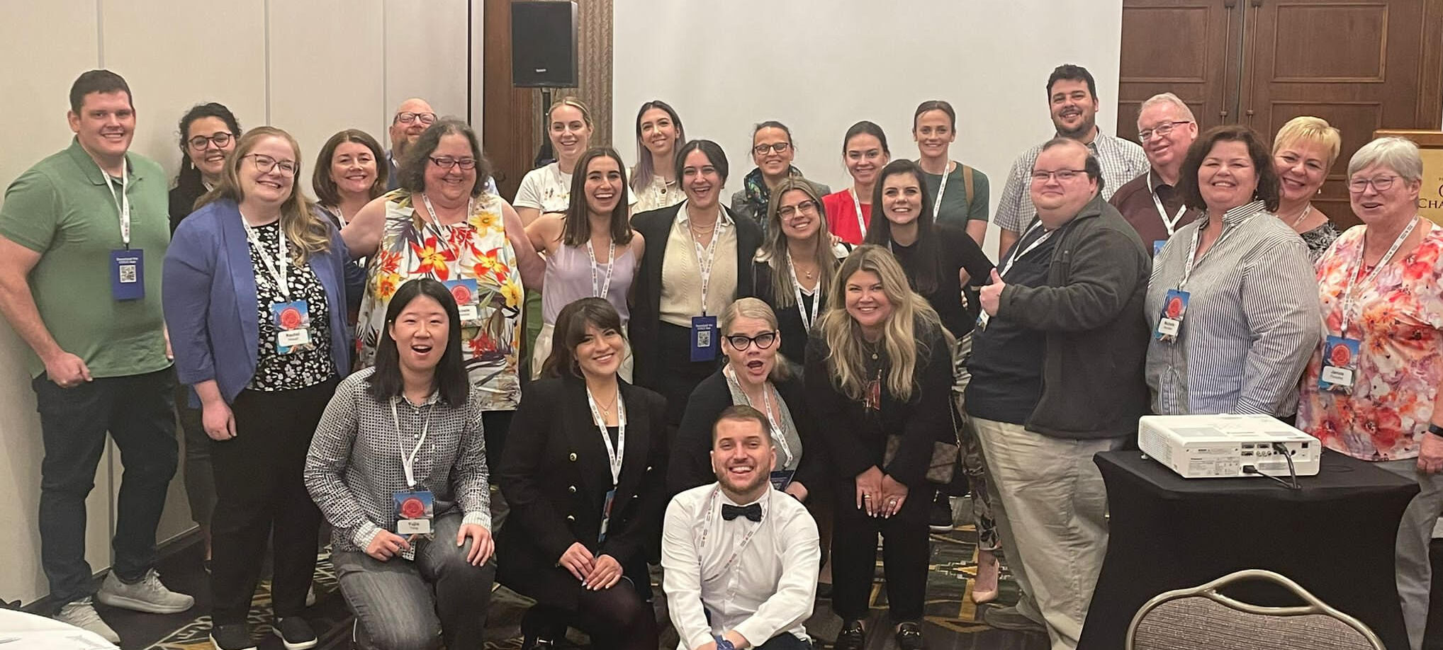 Group of smiling people posing for a photo in a conference room, some seated, others standing, all wearing conference badges.