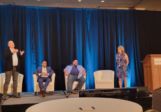 Four adults on a stage at a conference: one woman and three men, interacting in front of an audience, with blue curtains in the background.