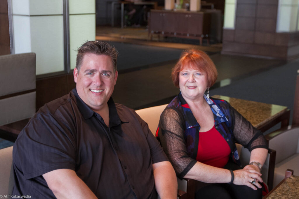 A middle-aged man and an older woman sitting at a cafe, smiling at the camera. both are dressed in casual attire.