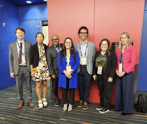 Group of seven professionals smiling for a photo at a conference, standing in front of a red wall.