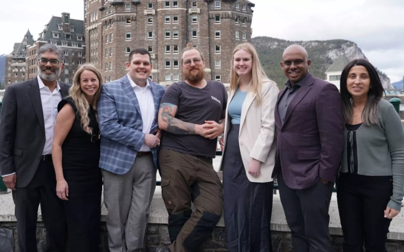 Seven people posing together in front of a historic stone building with mountains in the background.