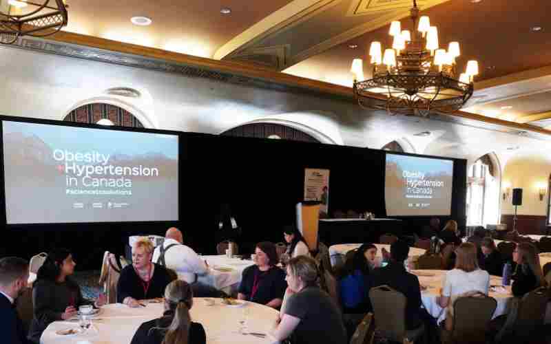 Conference attendees seated at round tables in a well-lit room, listening to a presentation about "obesity & hypertension in canada" displayed on a large screen.