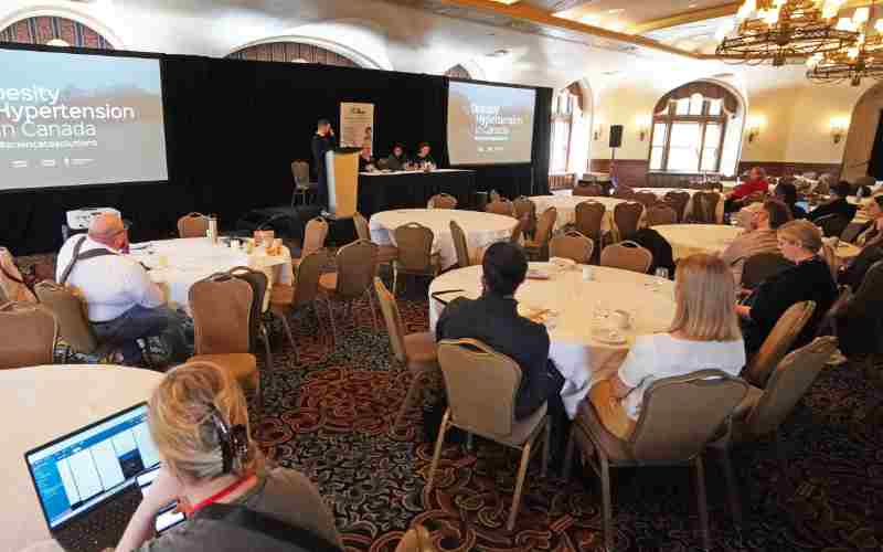 Audience seated in a conference hall listening to a speaker at a podium discussing obesity and hypertension in canada.