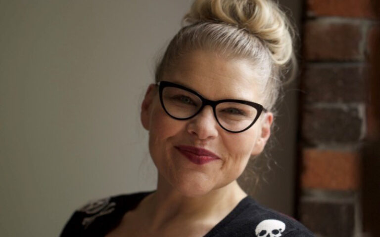 A person with blonde hair in a bun, wearing black-framed glasses and a black top with skull designs, smiles in front of a brick wall.