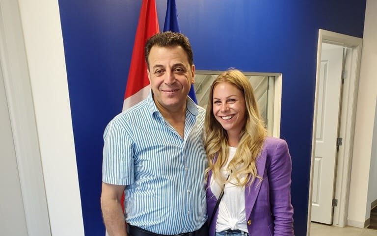 Two people standing side by side in front of a blue wall with a doorway and flags in the background. Both are smiling and wearing casual clothing.