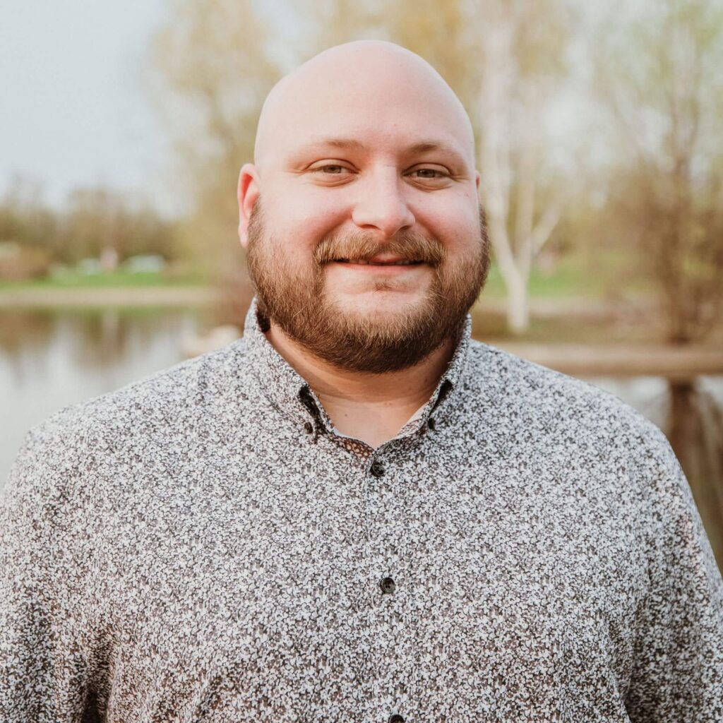 A man with a beard stands in front of a lake in a park smiling at the camera