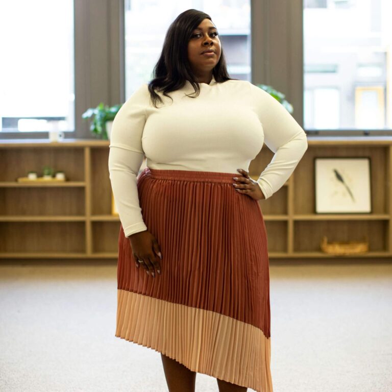 A Black woman in a cream sweater and red skirt stands in an office with one hand on her hip.