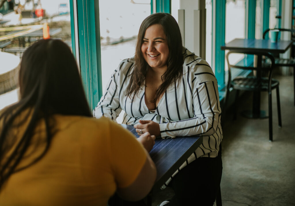 Two women are sitting in a cafe talking. One woman is facing the camera and the other has their back to the camera.