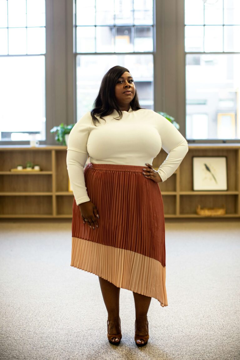 A woman in a white sweater and red skirt is standing in an office with one hand on her hip.