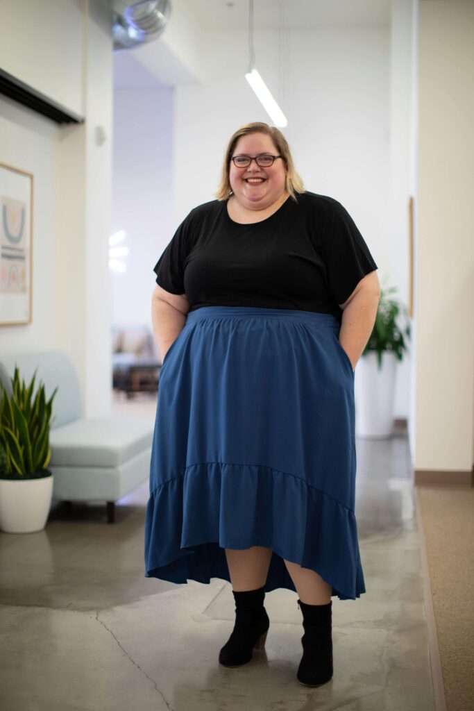 A woman wearing a black shirt and blue skirt stands and smiles at the camera