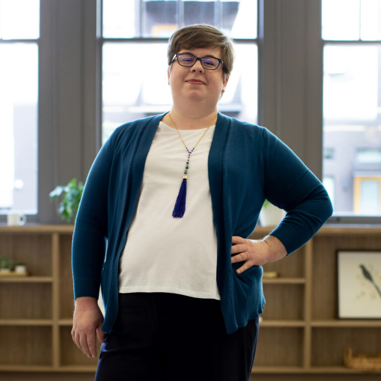 A woman with short blonde hair is standing looking at the camera with one hand on her hip. She is wearing a white blouse and teal sweater.