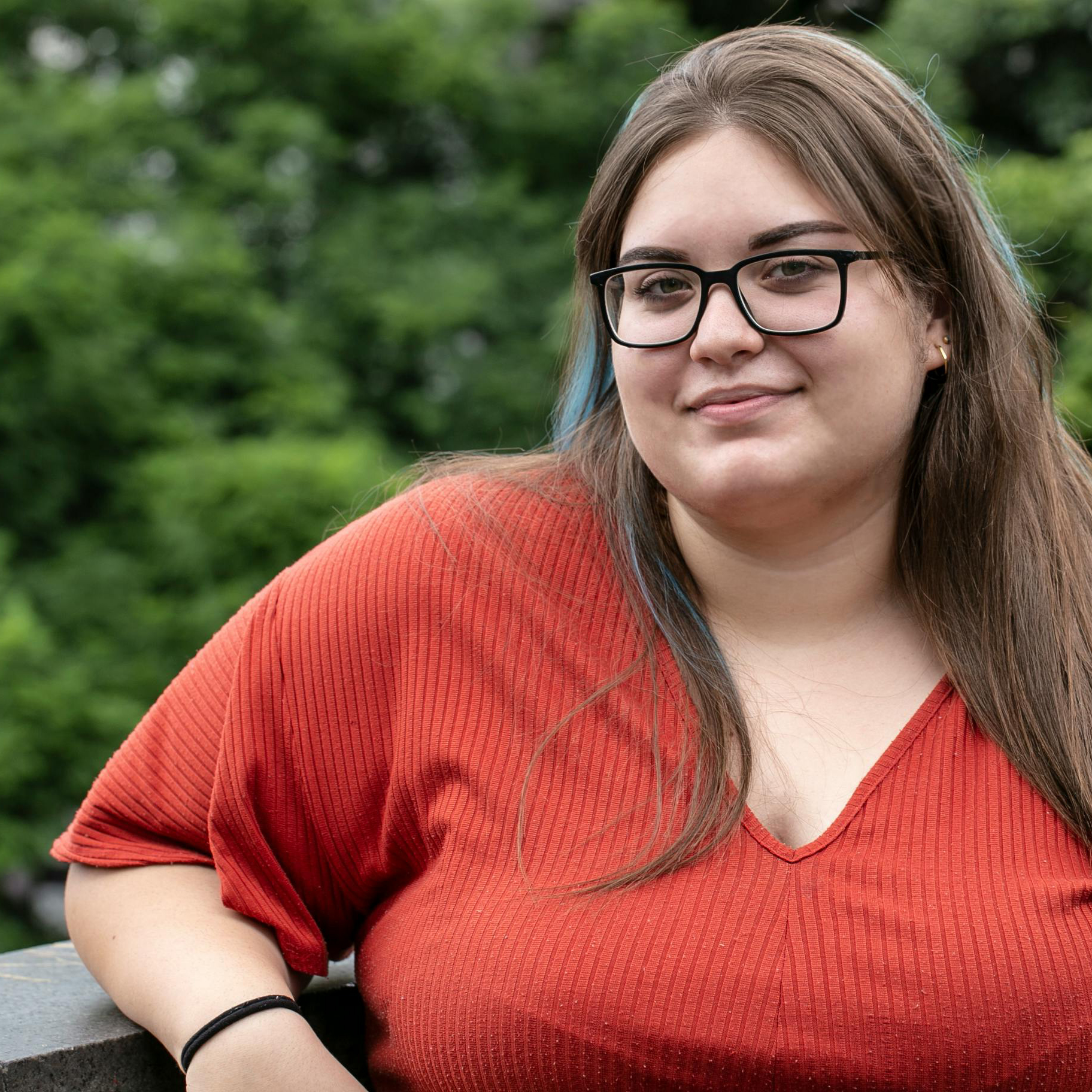 A woman with brown hair wearing glasses and a red shirt is outside with green trees behind her.