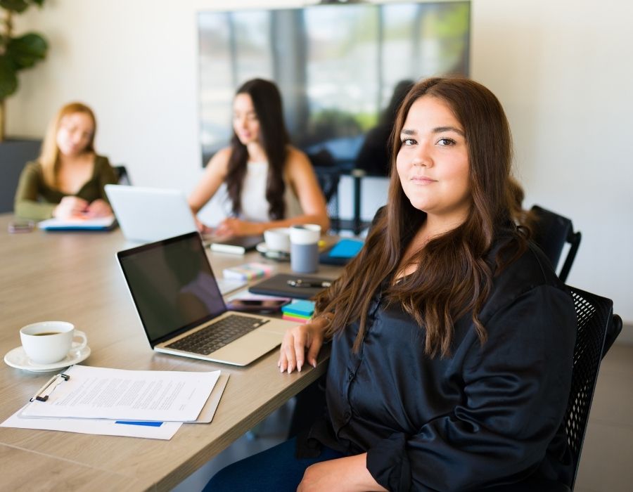 a woman with obesity sits at a conference room desk with her laptop in front of her.