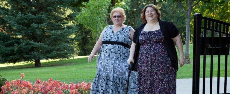 Two women wearing floral dresses walking through a park. One woman is using a cane for assistance.