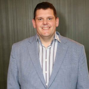 Dr. Ian Patton, PhD stands in front of a green background wearing a blue blazer and striped shirt.
