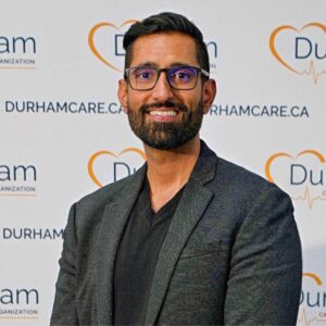 Dr. Rishi Handa wearing a grey suit standing in front of a background that says Durham Care Clinic + Pharmacy