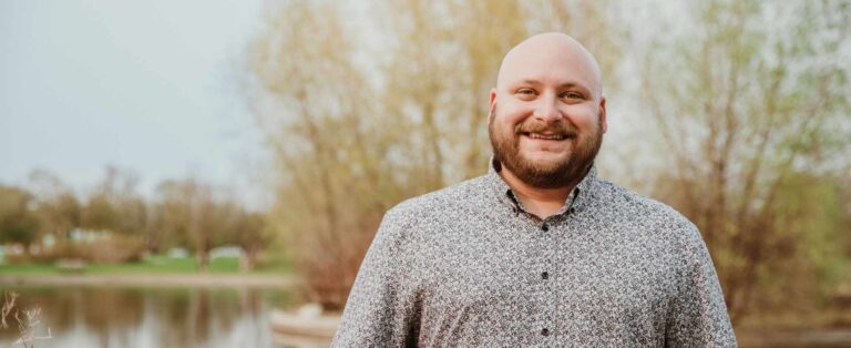 a man smiling at the camera outside in front of a small creek with trees