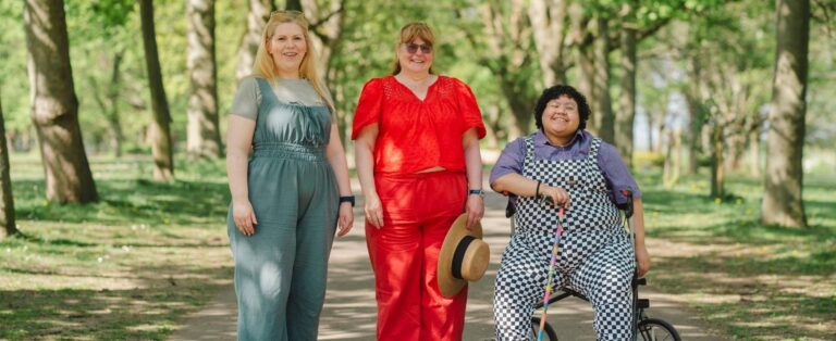 Three women in larger bodies standing outside in a park. They are all wearing colorful clothes and smiling at the camera.