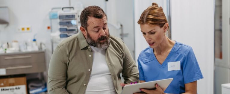 a man with a beard sitting in a doctor's office speaking with a clinician. They are looking at a tablet.