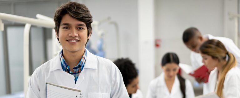 A medical student wearing a white coat and holding a notebook smiles at the camera. A group of medical students are gathered in discussion behind him.