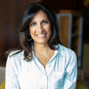 Dr. Shahebina Walji is wearing a white button-up shirt, smiling at the camera. Her background is blurred behind her