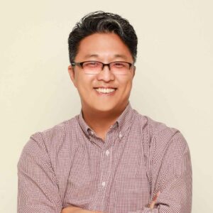Dr. James Kim is wearing a red checkered button up shirt against a beige background. He is wearing glasses and smiling at the camera.