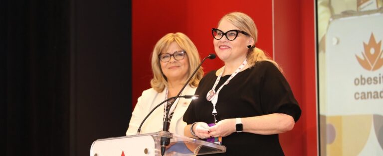 Lisa Schaffer, Executive Director of Obesity Canada, and Meghan Belland, Executive Director of Primary Care Alberta, stand on stage at an Obesity Canada conference. There is a red backdrop behind them.
