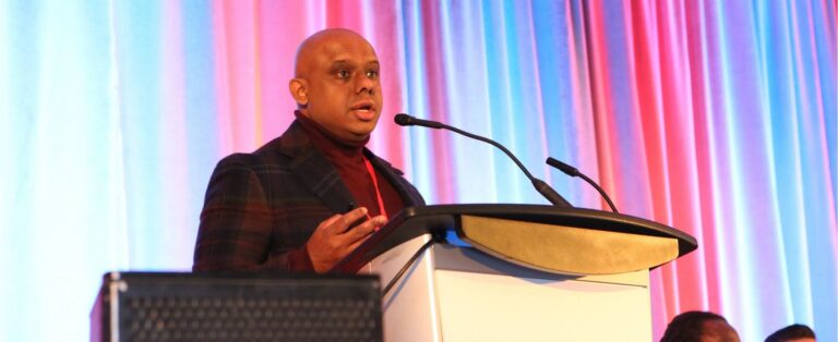 Dr. Sanjeev Sockalingam stands speaking at a podium with a coloured drape background.