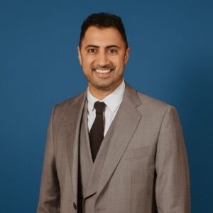 Marc Bains from Heartlife Foundation smiles at the camera while wearing a grey suit and black tie against a blue background.