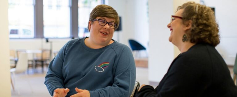 Two people sit facing each other in a bright indoor space, smiling and talking. One wears glasses and a blue sweatshirt with a small rainbow graphic, while the other wears glasses and a black top.