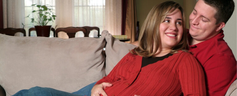 A pregnant person in a red cardigan sits on a couch, smiling at their partner as they relax together at home.